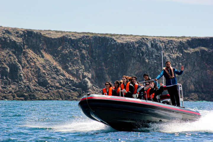 People on a speedboat wearing life jackets near rocky cliffs.