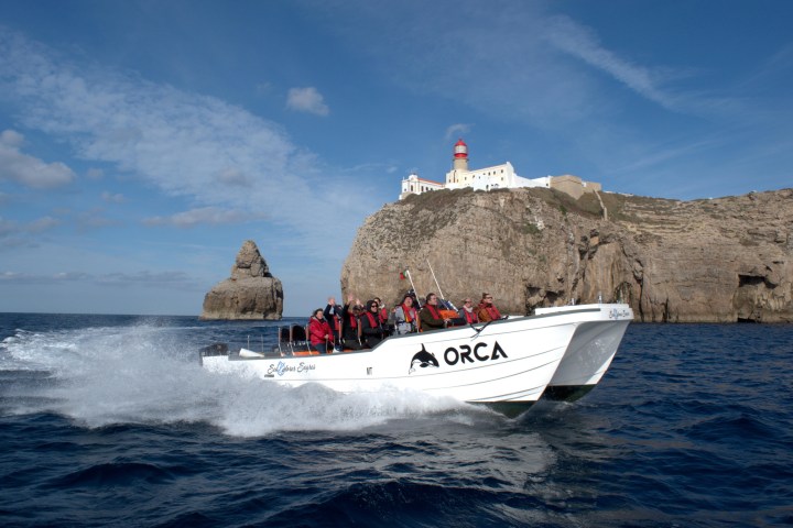 A speedboat named ORCA speeds near cliffs with a lighthouse on top under a blue sky.
