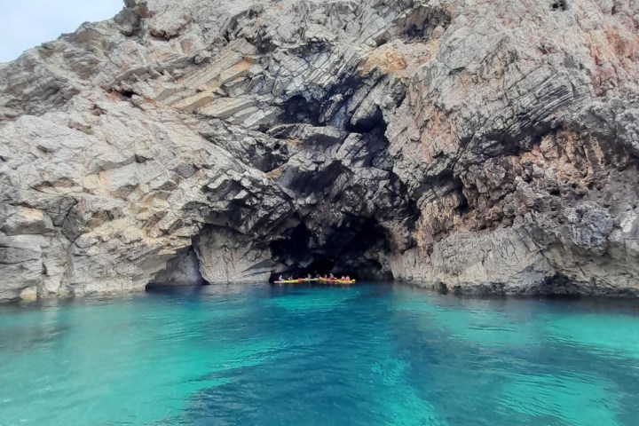 Rocky cliff with a sea cave and clear blue water, with kayakers inside the cave.