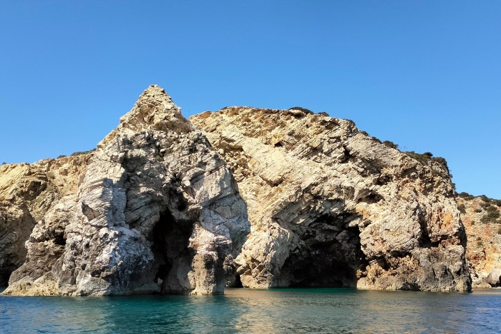 Rocky coastal cliff with caves under a clear blue sky, surrounded by calm sea water.