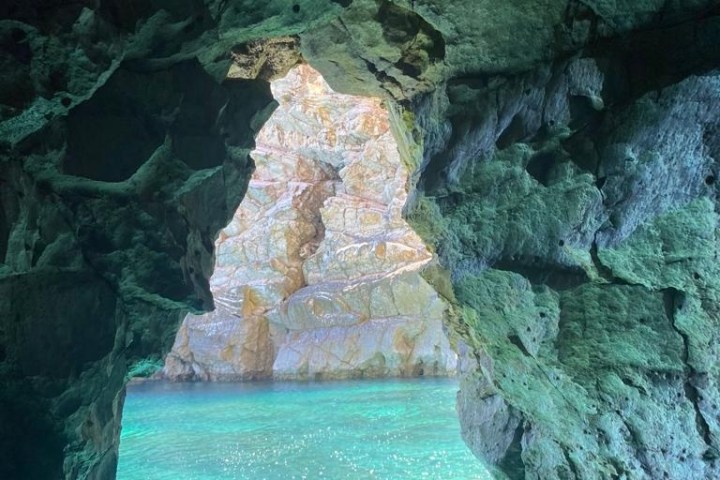 Boat entering a rocky cave with turquoise water and a person holding a camera.