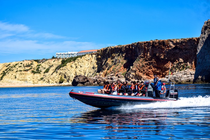 Speedboat with people in life vests near rocky coastline under clear sky.