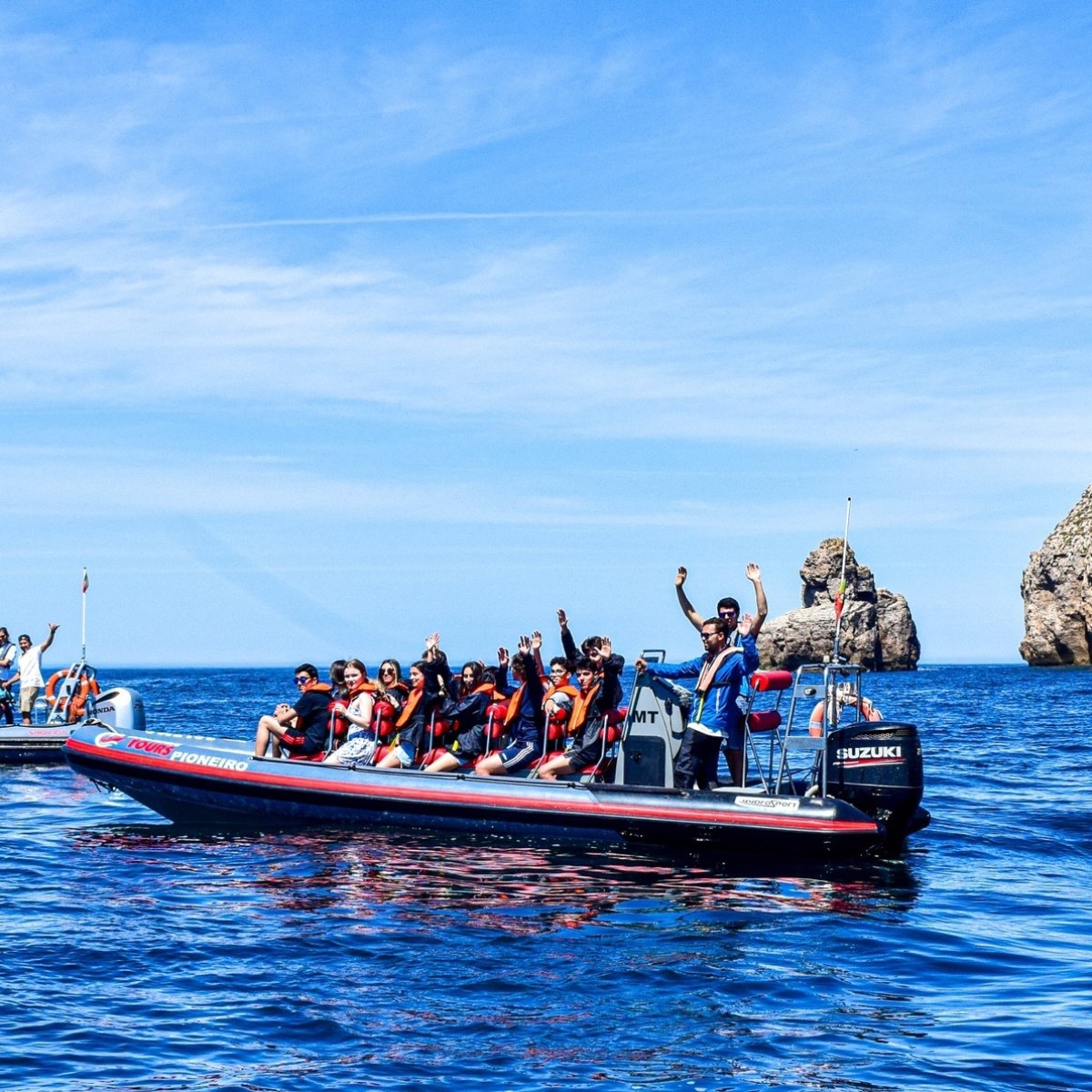 Two boats with people in life vests wave in the sea near a rocky cliff with a lighthouse.