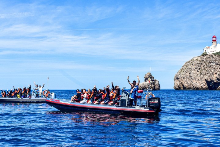 Two boats with people in life vests wave in the sea near a rocky cliff with a lighthouse.