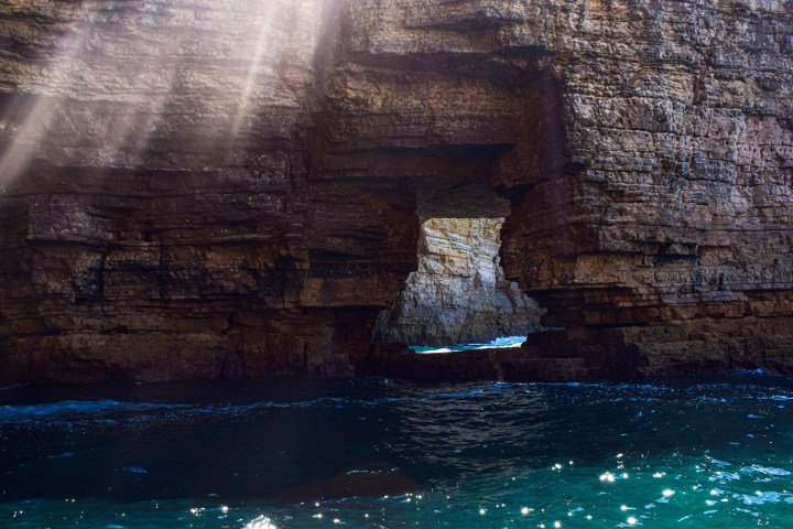 Sunlight streaming through a rocky archway over blue ocean water.