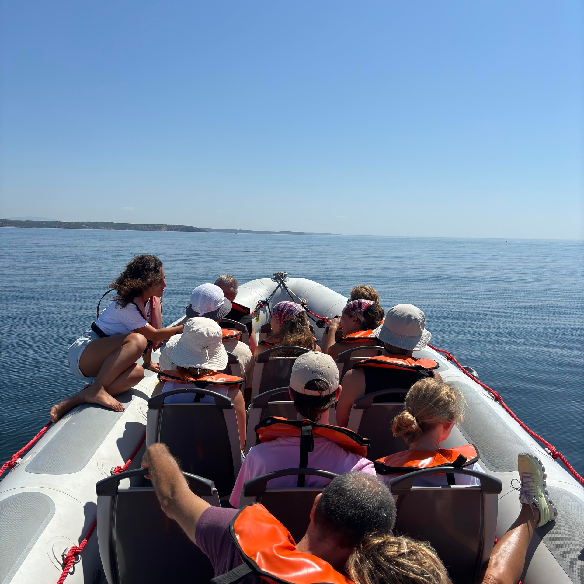People in life vests sit on a boat in calm waters under a clear blue sky.