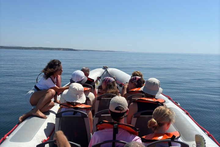 People in life vests sit on a boat in calm waters under a clear blue sky.