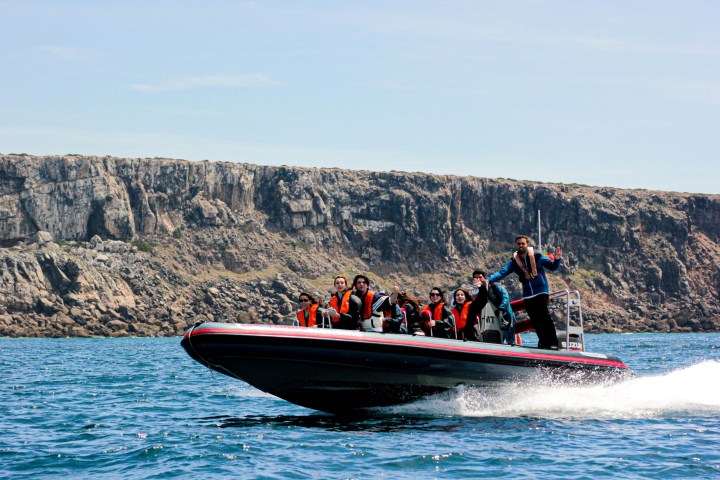 People in life jackets on a speedboat near rocky cliffs.