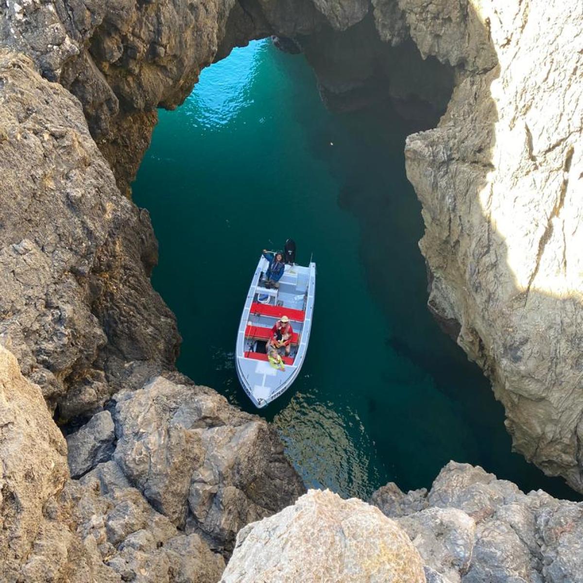 Boat with two people in a rocky cove with blue water.