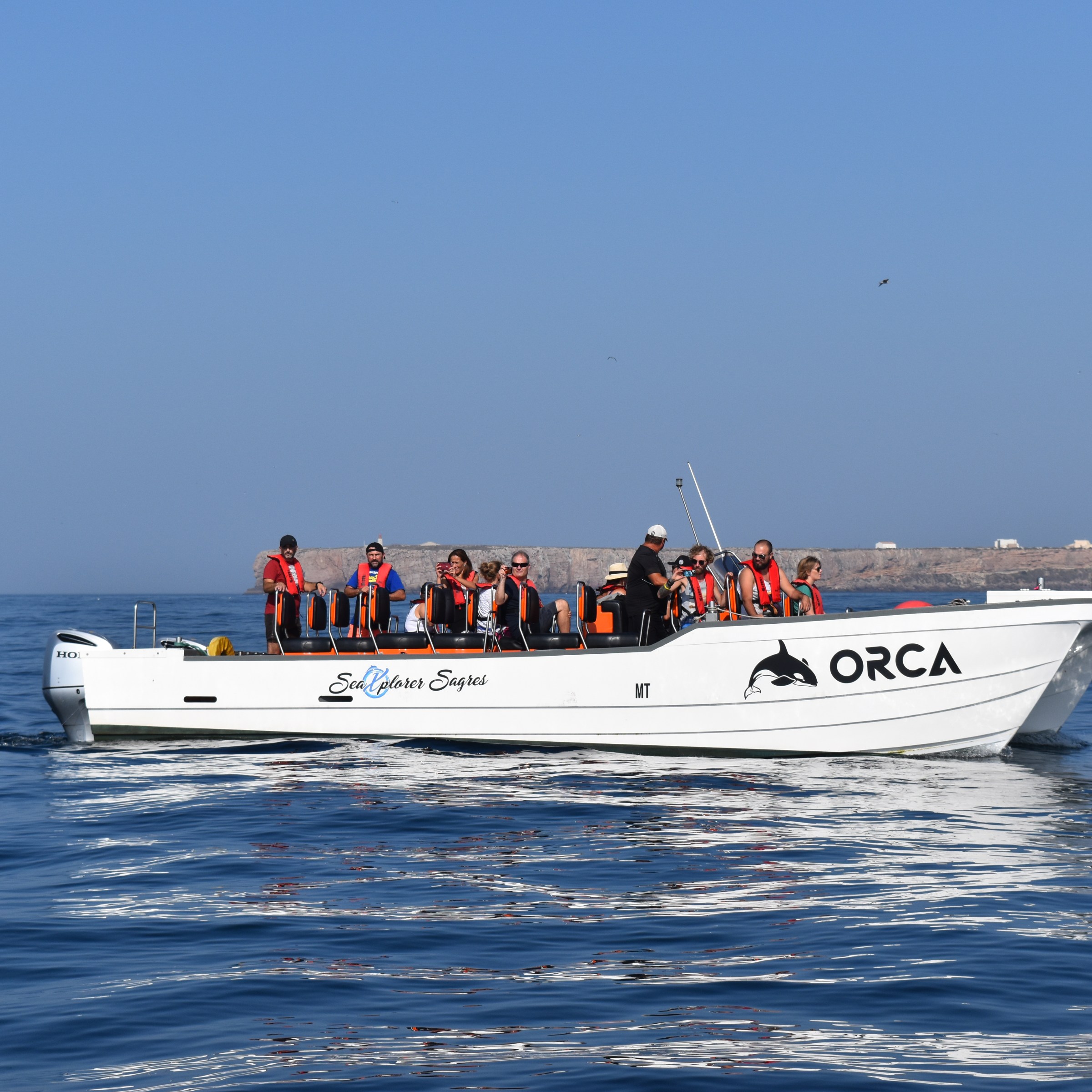 People on a white boat labeled 'ORCA' on calm sea under clear blue sky.